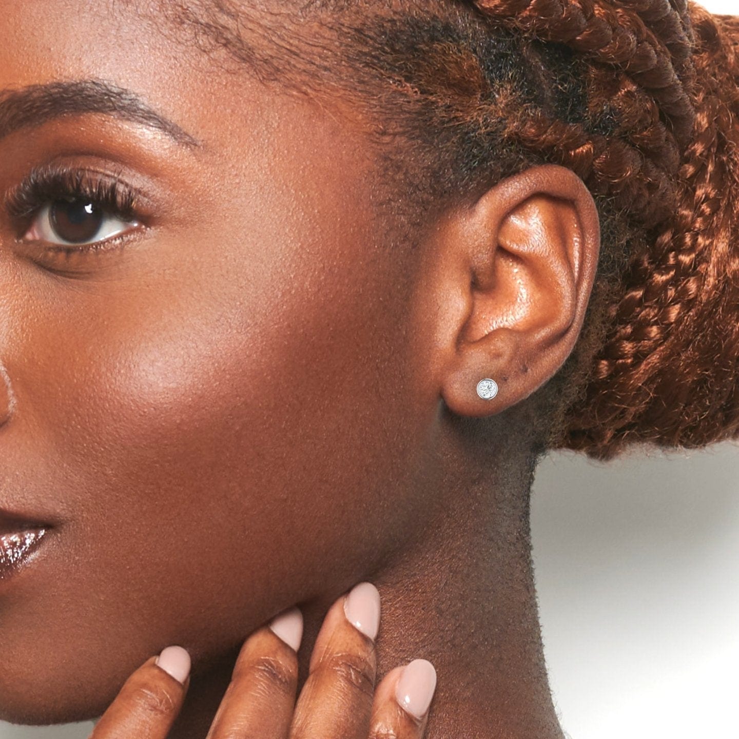 Close-up of a woman with glowing brown skin, braided hair, and manicured nails touching her chin. She wears the Ishanvi Lab Diamond Earrings - Bezel Set, paired with natural makeup against a white background.