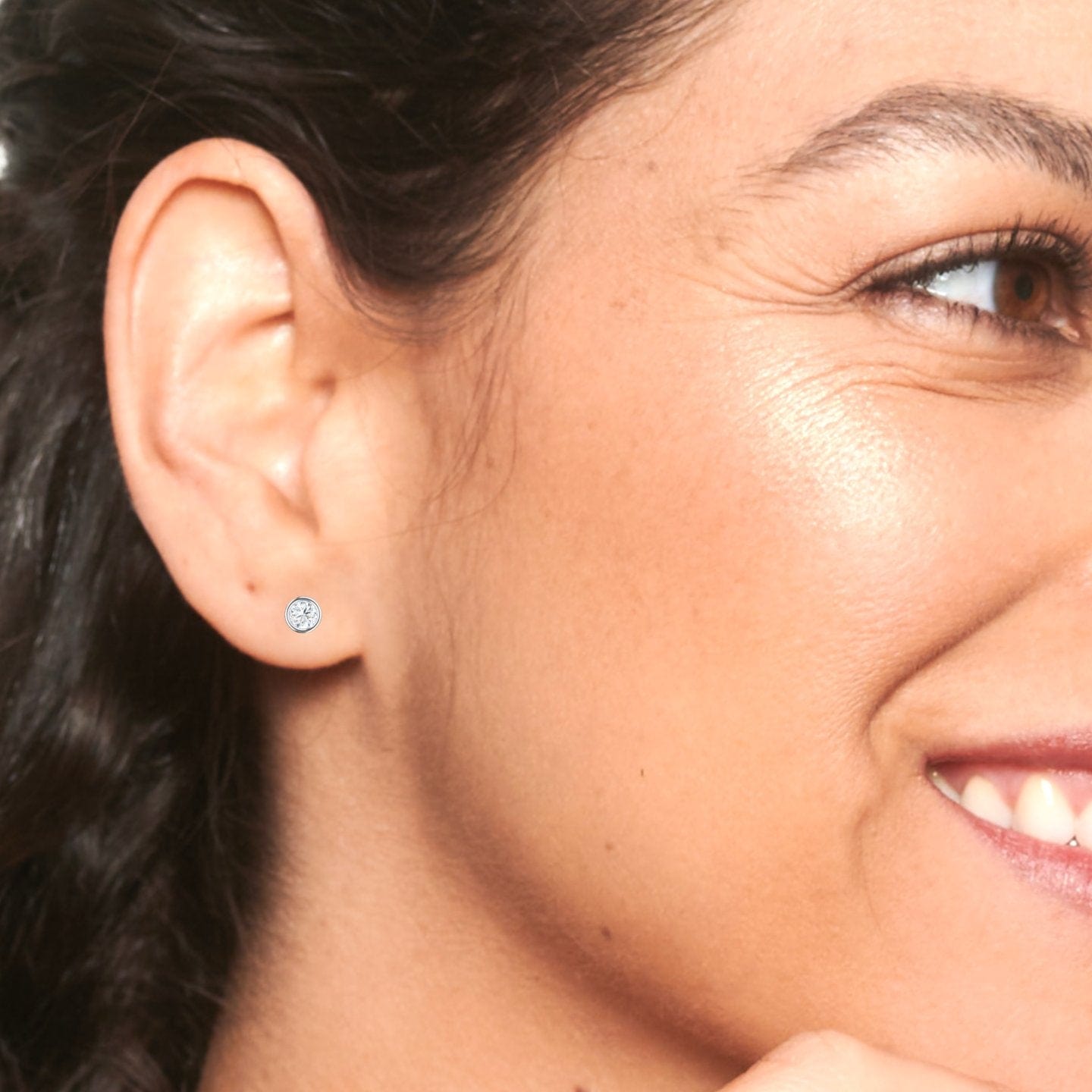 Close-up of a woman wearing the Lab Diamond Earrings - Bezel Set - Ishanvi, featuring a small round diamond stud on her ear. She smiles softly, with smooth skin and dark hair braided to the side.