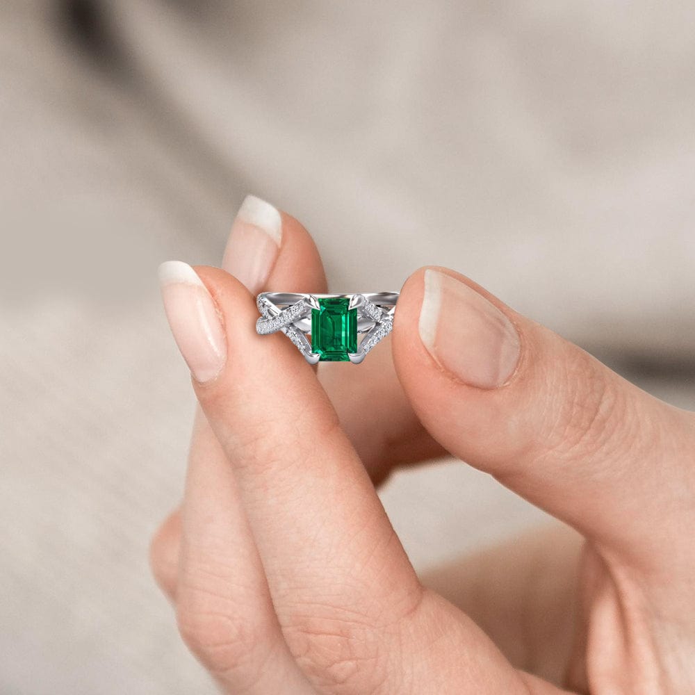Hand holding a diamond ring with a green emerald gemstone against a neutral background