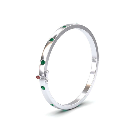 The Emerald Bezel-Set Bracelet - Flavia is a polished silver bangle with evenly spaced green gemstones, a small round red accent, and a clasp closure, shown against a plain white background.