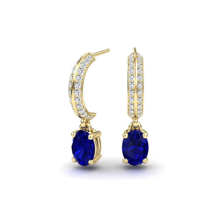 The Blue Sapphire & Diamond Huggie Drop Earrings – Serena feature oval blue sapphires suspended from gold hoops set with a row of small white diamonds, shown against a white background.