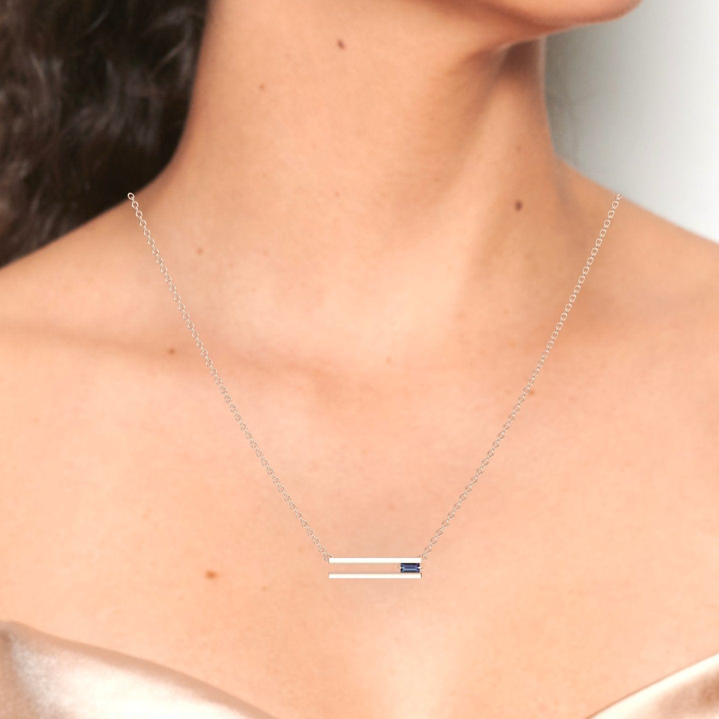 A woman wears the Cecilia Lab Grown Alexandrite Bar Necklace, featuring two parallel silver bars—one accented with a blue gemstone. She is dressed in a light-colored, off-shoulder top.