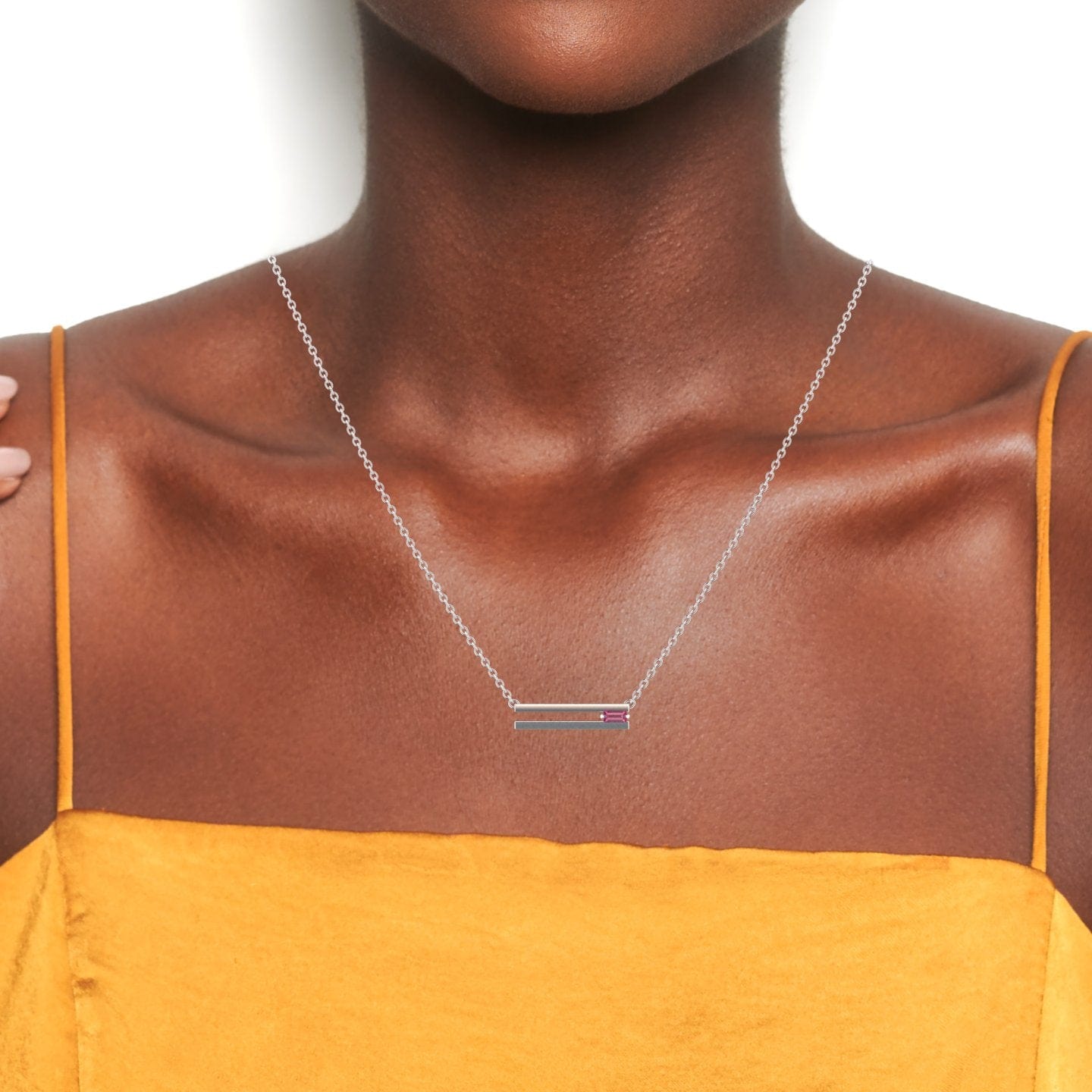 A person wearing the Pink Tourmaline Bar Necklace - Cecilia, a thin silver chain with a rectangular pendant adorned with pink stones, styled with a mustard yellow sleeveless top against a white background.
