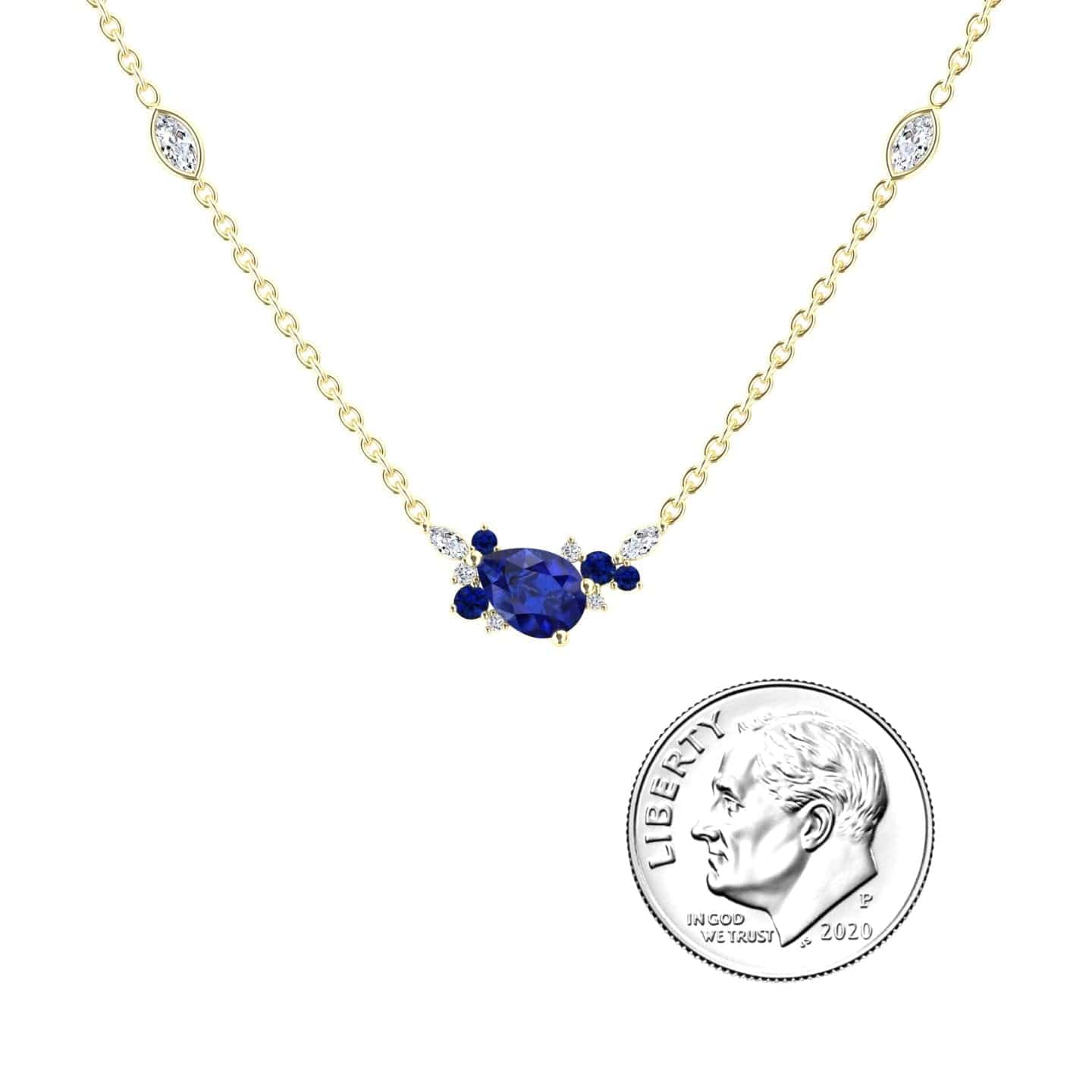 The Blue Sapphire & Diamond Bouquet Necklace - Daria features a gold chain and a central blue oval gemstone encircled by clear and blue stones, shown beside a U.S. dime for size comparison.