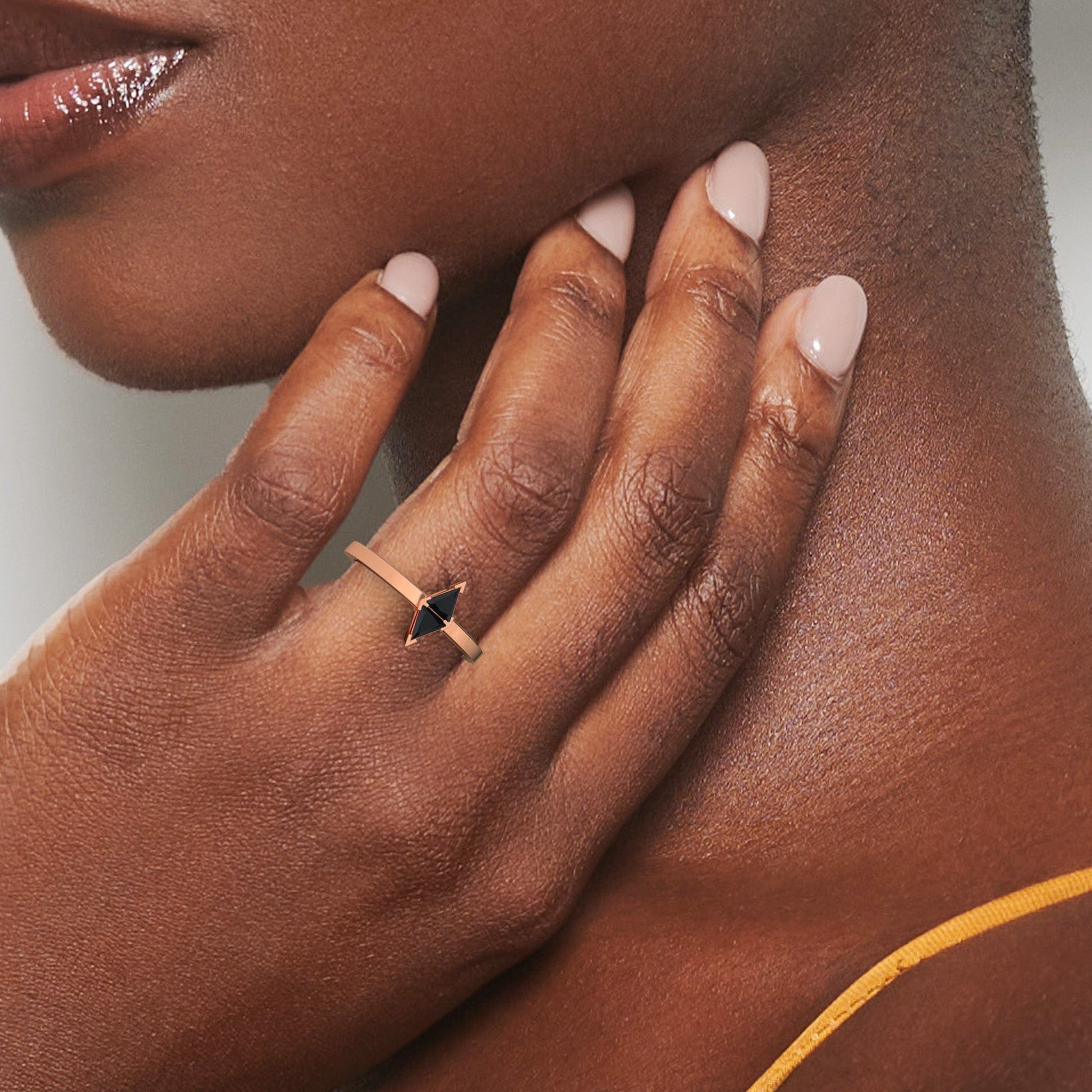 A close-up of a person with dark skin wearing the Black Diamond Triangle Statement Ring - Shalini, featuring a bold black gemstone, as they touch their neck. They have nude nail polish and wear a yellow strap top.