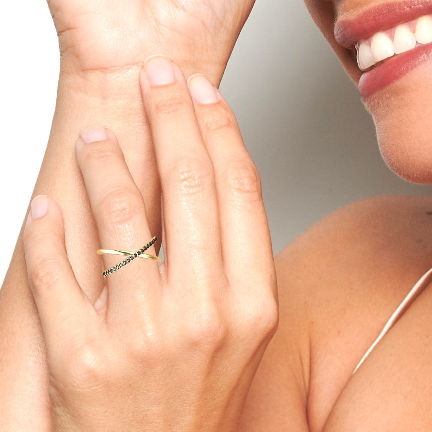 A smiling person touches their face, displaying a close-up of their hand adorned with the Black Diamond Criss-Cross Pave Band Ring - Trisha, featuring one gold band and another set with sparkling black stones.