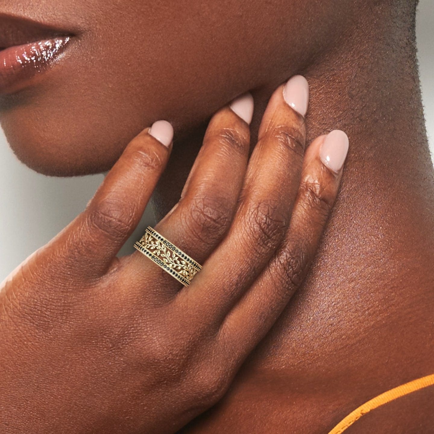 A womans hand with pale pink nails rests on her neck, highlighting the Black Diamond Vine Motif Eternity Band - Casey, a gold ring with intricate vine details. Her smooth, rich-toned skin and glossy lips add to the elegance.
