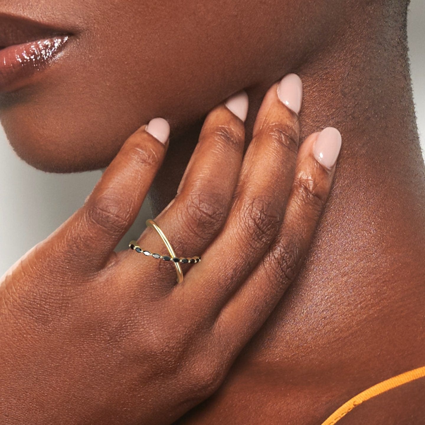 A close-up of a hand with light pink nails rests on a neck, highlighting the Gracie Black Diamond Crossover Ring—a gold band with delicate black stones—against smooth skin and a softly blurred background.