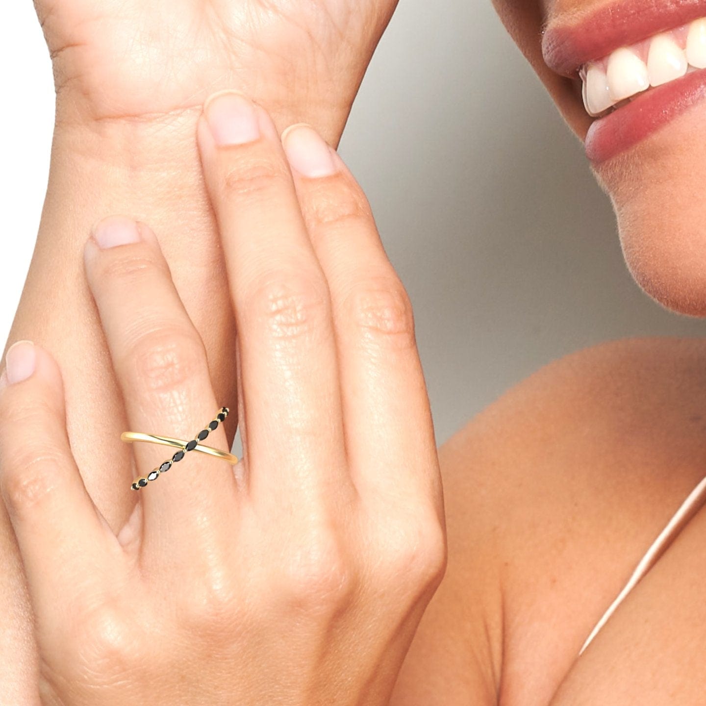 A close-up shows a smiling person wearing the Black Diamond Crossover Ring - Gracie on their finger, with black stones set in gold, as their other hand gently touches the ring. Lips and part of a shoulder are also visible.