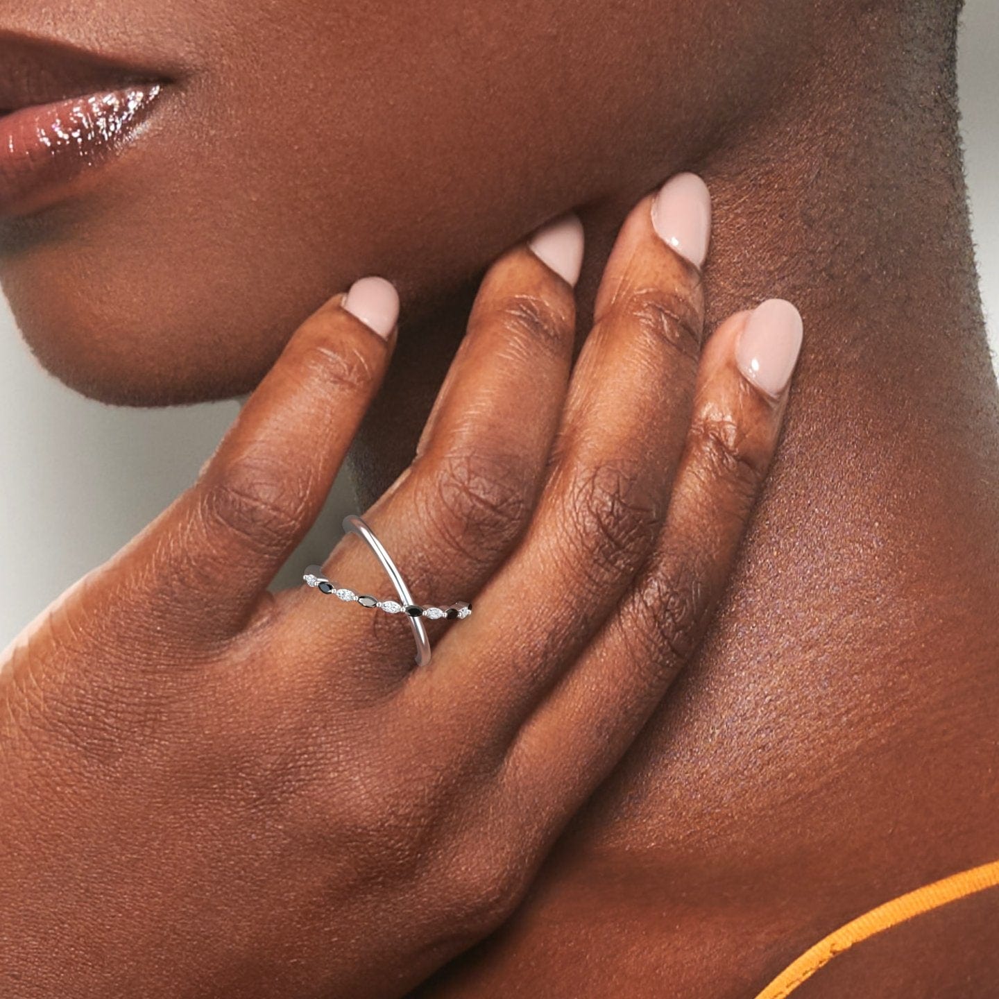 A close-up shows a hand with nude polished nails gently touching a woman’s neck, elegantly displaying the Gracie Black & White Diamond Crossover Ring. The delicate silver band and sparkling gemstones stand out against the softly blurred background.