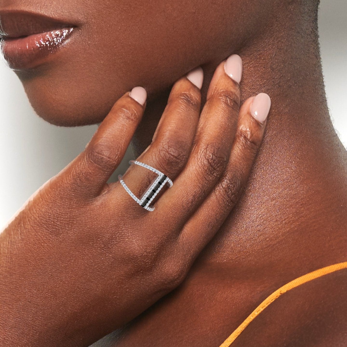 A close-up of a womans hand with nude nails touches her face, highlighting the sparkling Luci Black & White Diamond Double Band Open Cuff Ring. She wears a yellow strap and has warm, smooth skin.