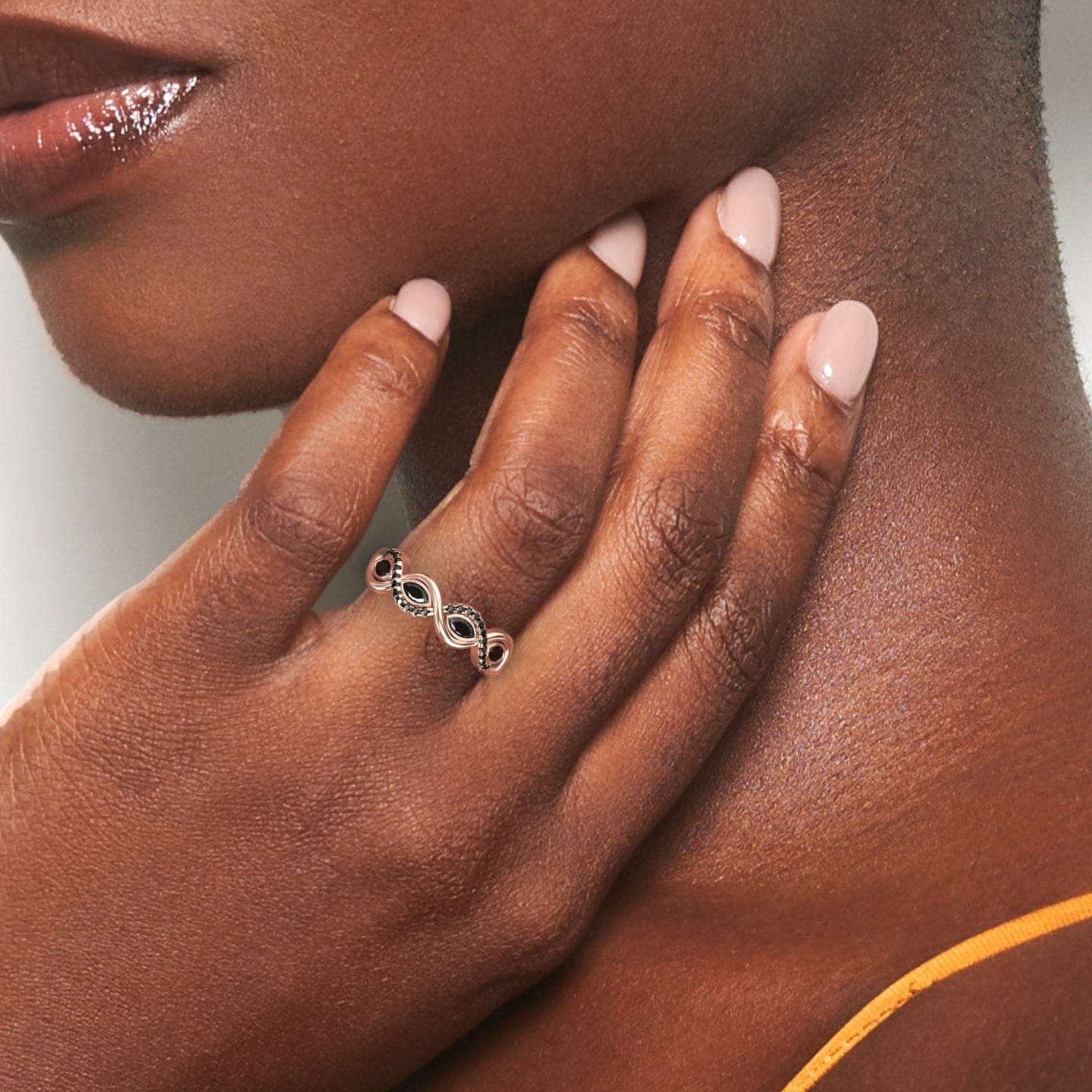 Close-up of a womans smooth dark hand with nude polish gently touching her neck, highlighting the Black Diamond Infinity Twist Band - Selma on her finger. She wears an orange strap and the silver ring sparkles with small black stones.