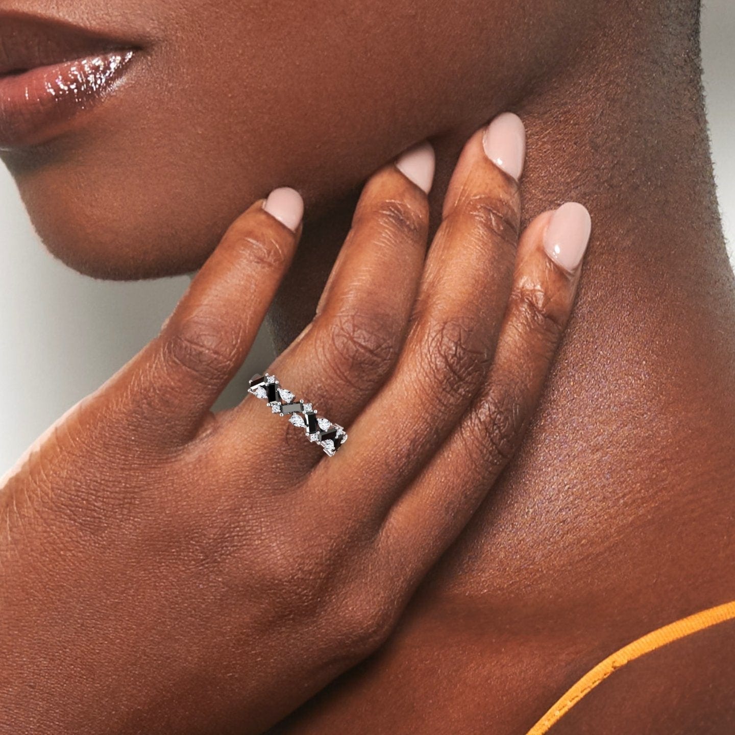 A close-up of a dark-skinned woman’s hand with pale pink nails gently touching her neck, showcasing the Black & White Diamond Zigzag Eternity Band - Aimee on her finger while wearing an orange strap.