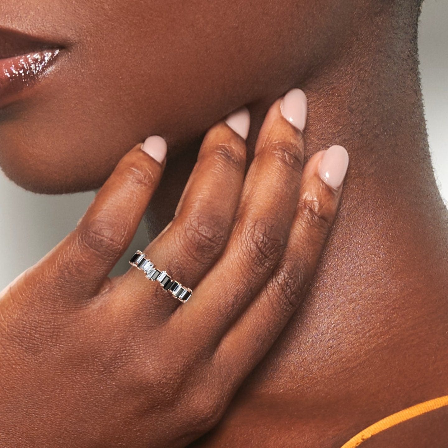 A close-up of a dark-skinned hand with light pink nails gently touching the neck, showcasing the Black & White Diamond Classic Eternity Band - Samantha, a silver ring with rectangular gems. The person is wearing a sleeveless top.