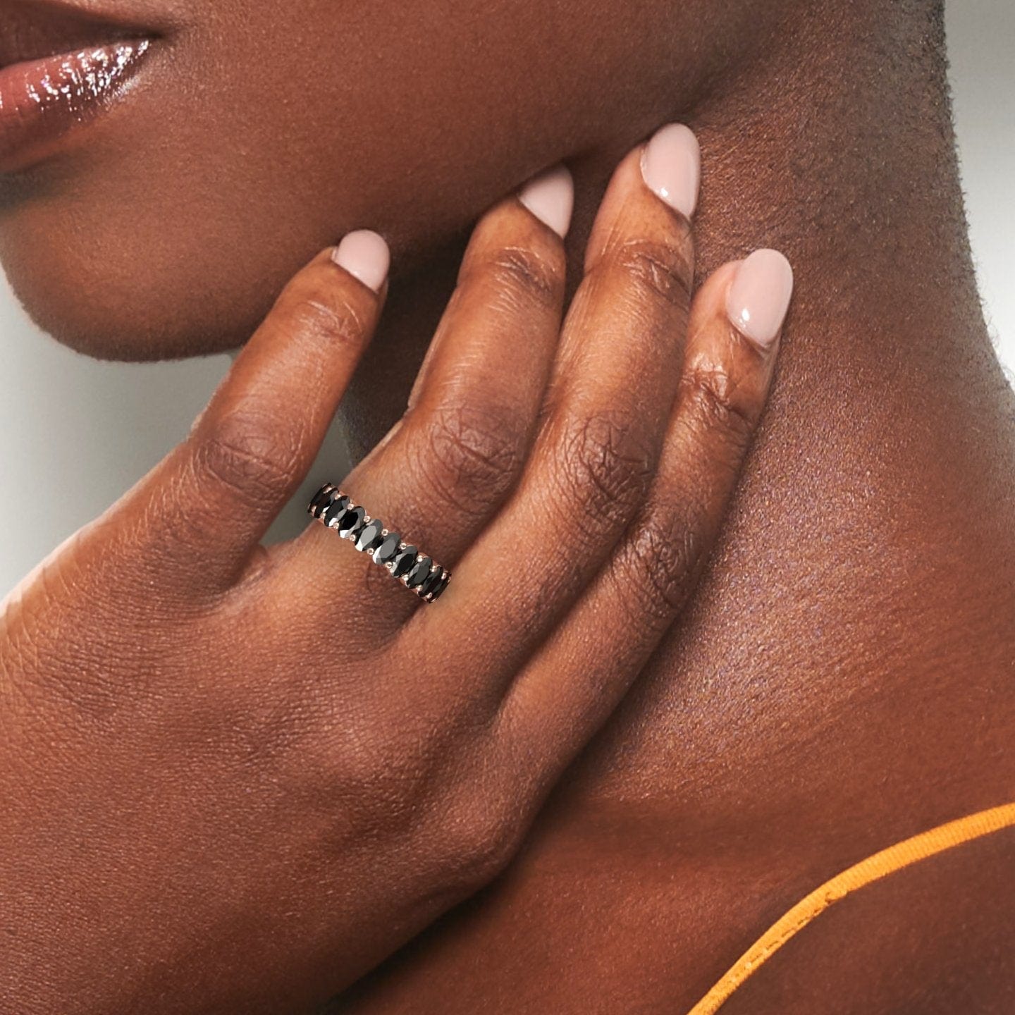 A womans hand with nude-pink nails gently touches her neck, showcasing the Black Diamond Full Eternity Band - Diana on her finger. She wears a thin yellow strap on her shoulder.