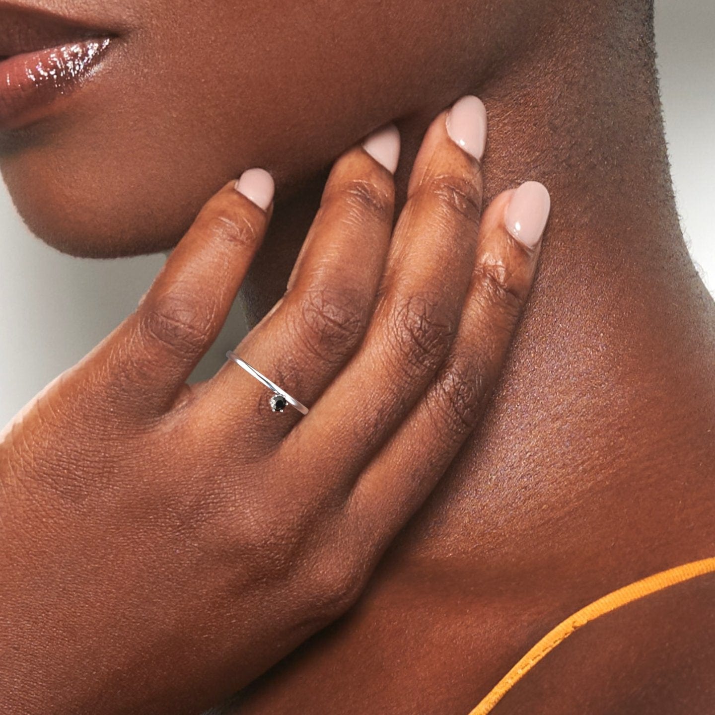 A womans hand with smooth dark skin and nude manicured nails rests on her neck, showcasing the Black Diamond Solitaire Petite Ring - Faria. She wears a yellow spaghetti strap top.