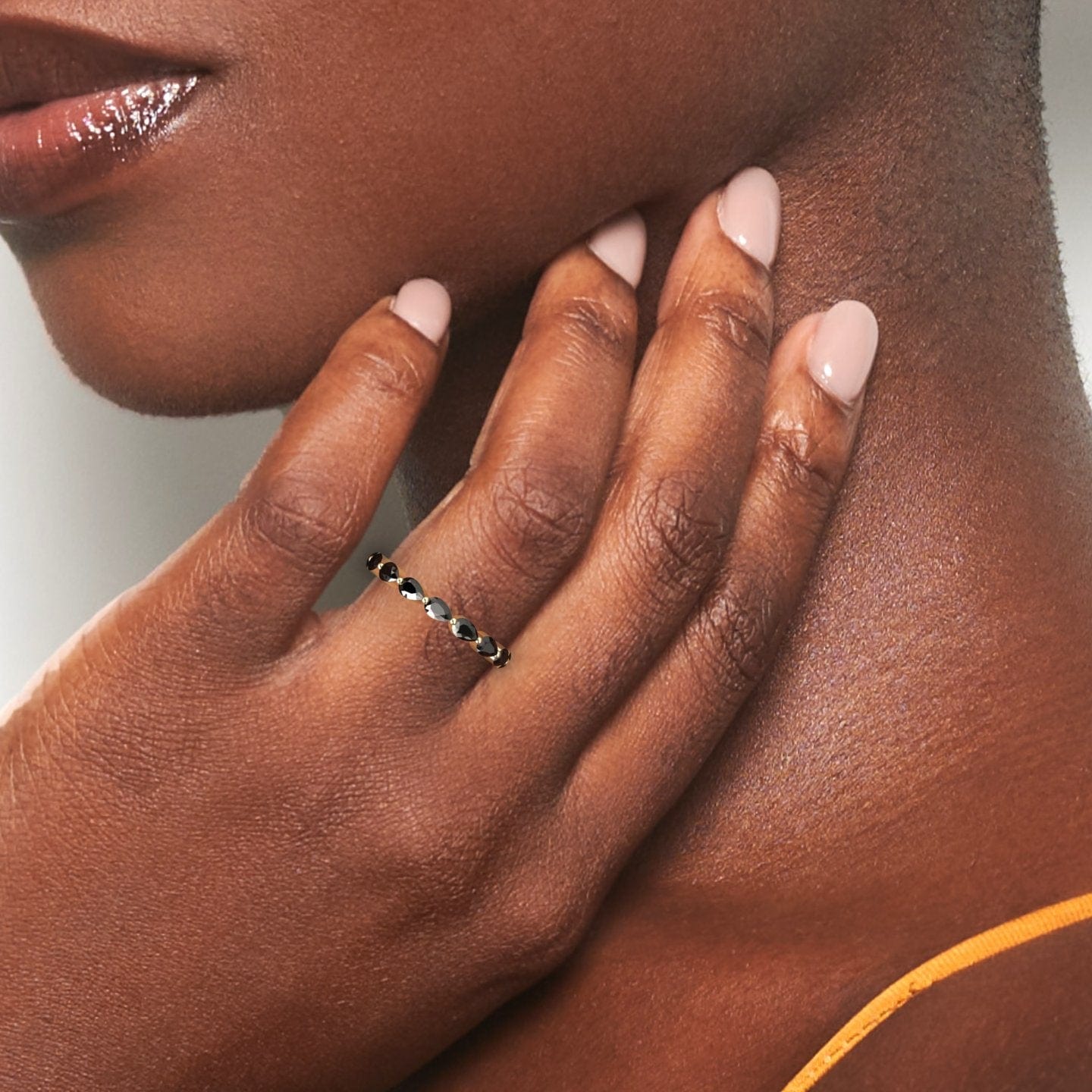 A woman with medium-dark skin touches her neck, showcasing the Kalyani Black Diamond Stackable Eternity Band on her manicured finger. A thin orange top strap is visible on her shoulder.