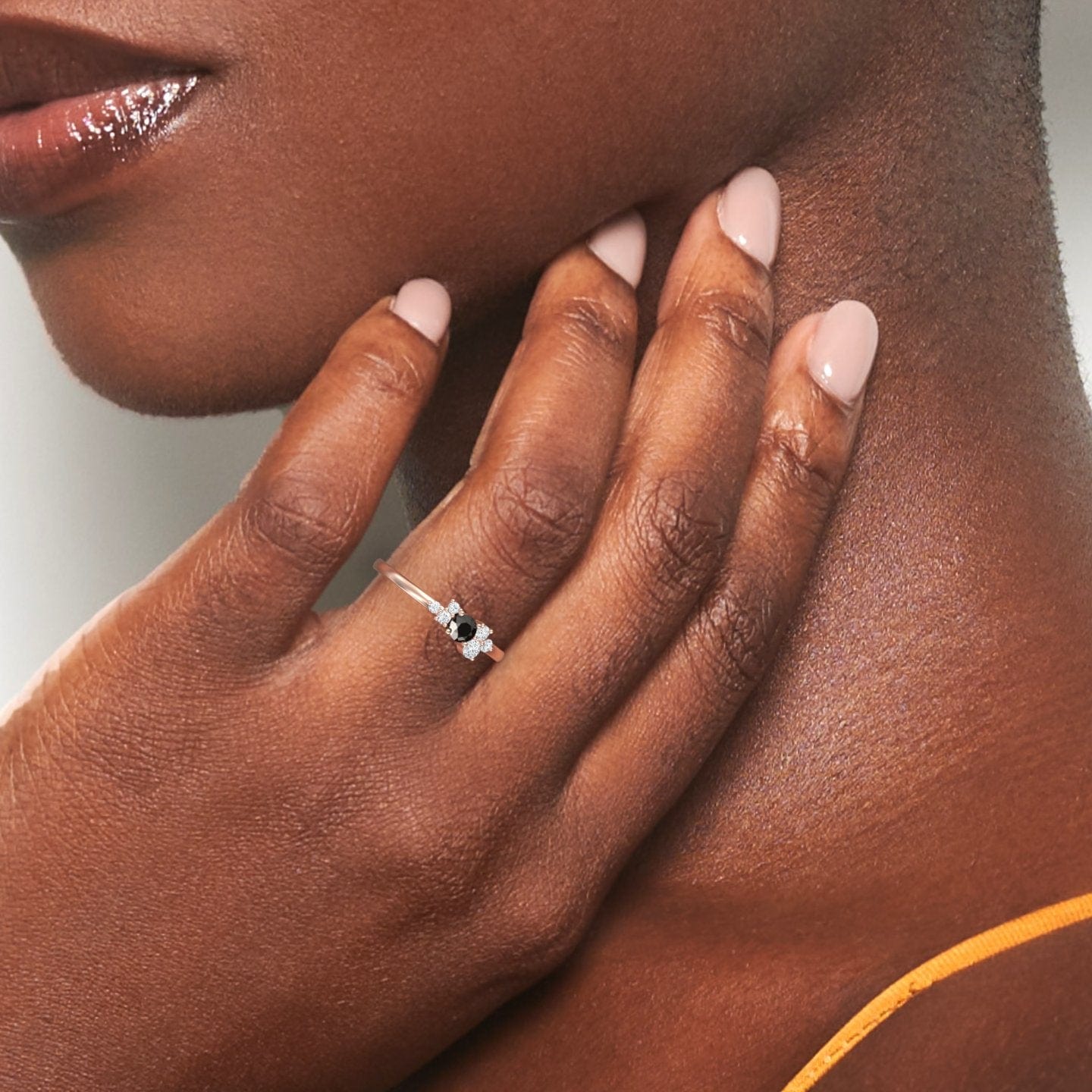 A close-up of a person with smooth brown skin and manicured fingers gently touching their neck, showcasing the Black & White Diamond Cluster Ring - Liana. The dark gemstone sparkles as they wear a thin orange strap.