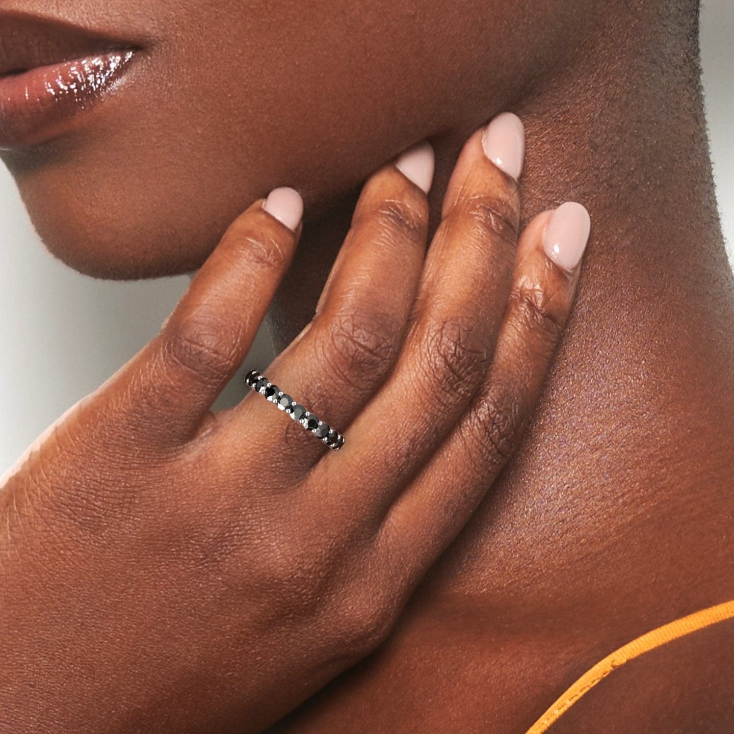 A close-up of a womanโs manicured hand touches her neck, displaying the Mahira Black Diamond Prong-Set Eternity Band on her finger. She has smooth skin and wears a thin orange strap.