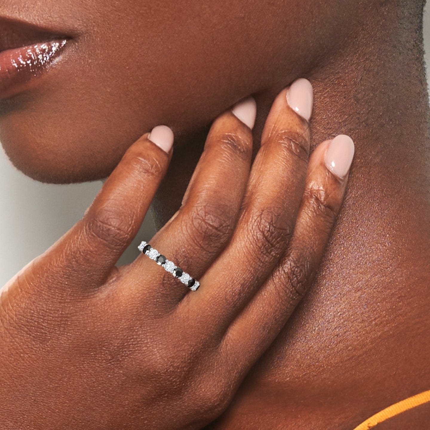 A womans manicured hand touches her neck, showcasing the Parveen Black & White Diamond Half Eternity Wedding Ring. She wears a coral-colored top and has smooth skin.