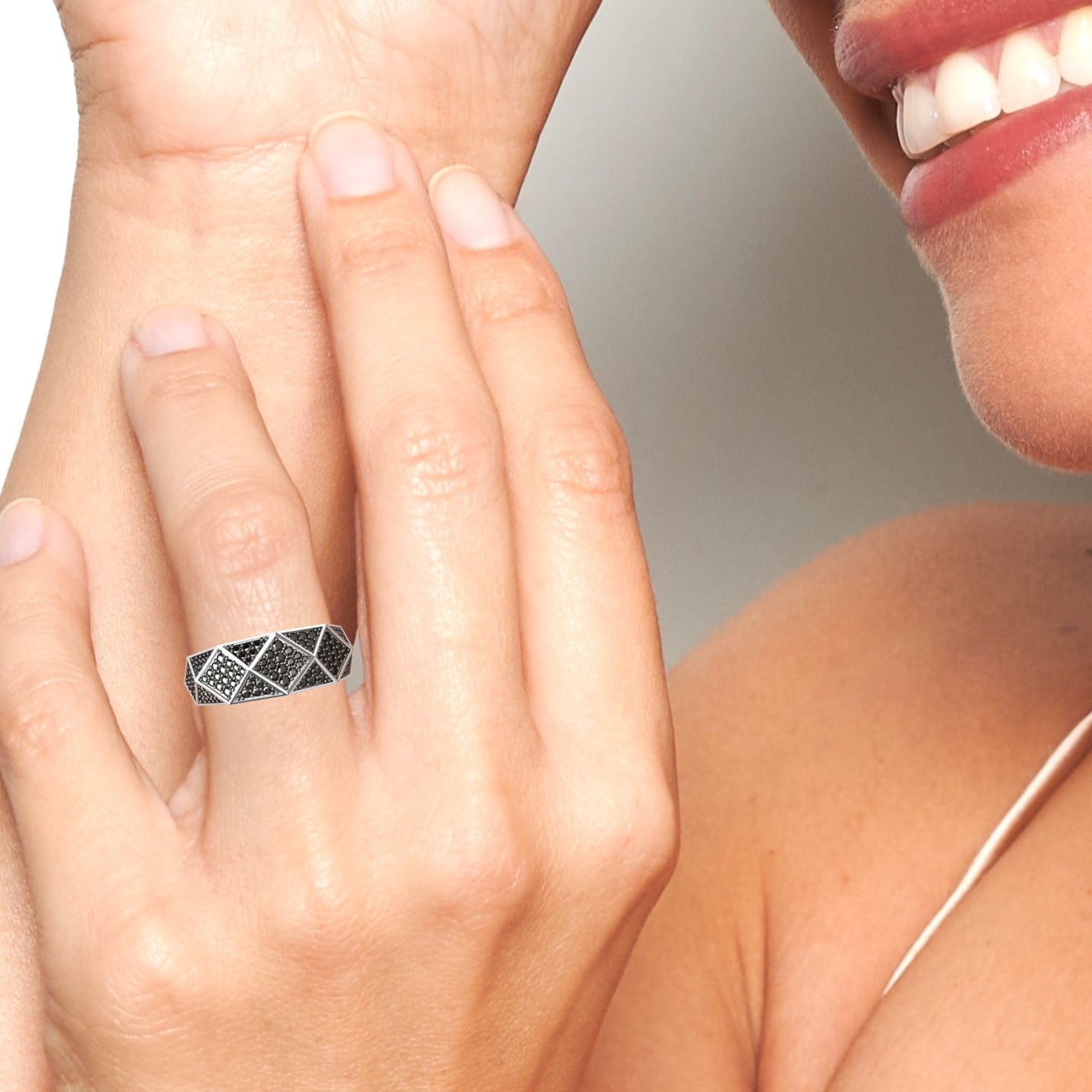 A person smiles and touches their face, wearing the Black Diamond Geometric Pave Set Statement Band - Blanca—a wide silver ring with a black and silver geometric design—on their middle finger against a plain, neutral background.