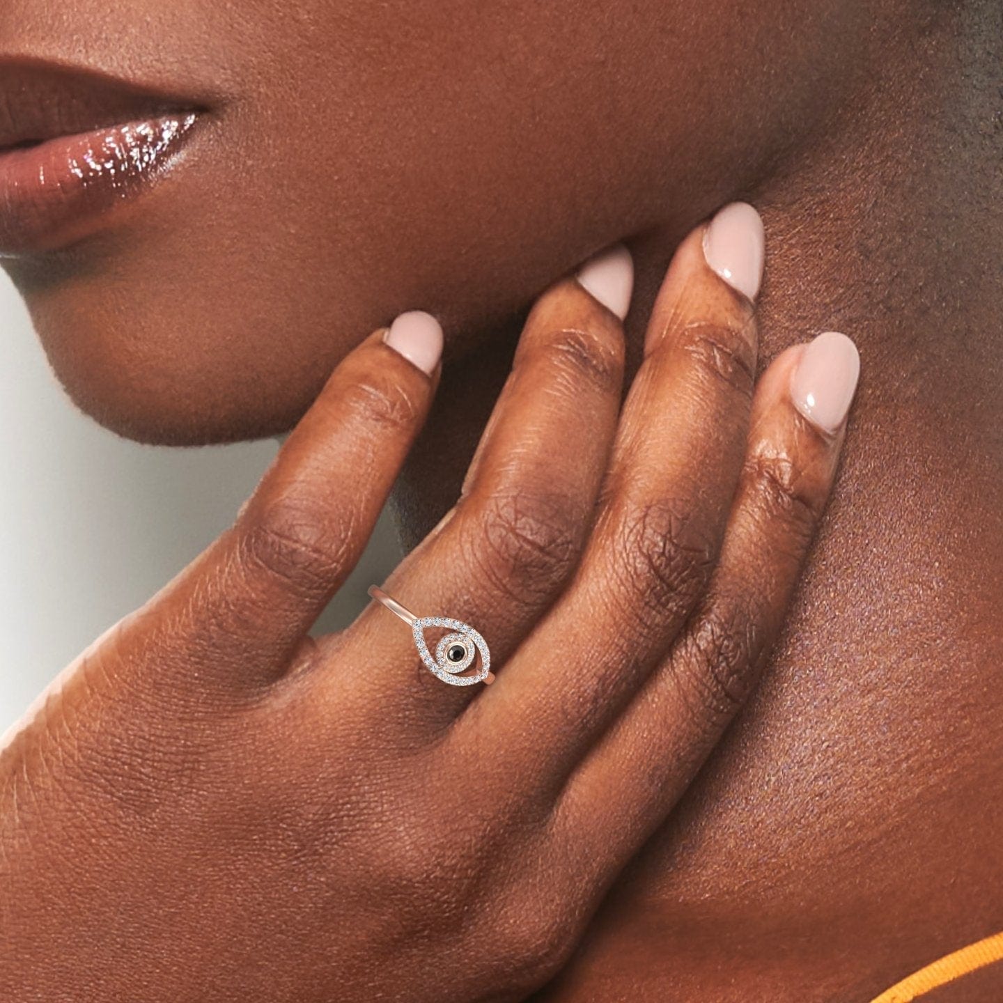 A womans pale pink nails rest on her neck, highlighting the Hazel Black & White Diamond Evil Eye Ring with a swirling design. Her lips are glossed and she wears a yellow top.
