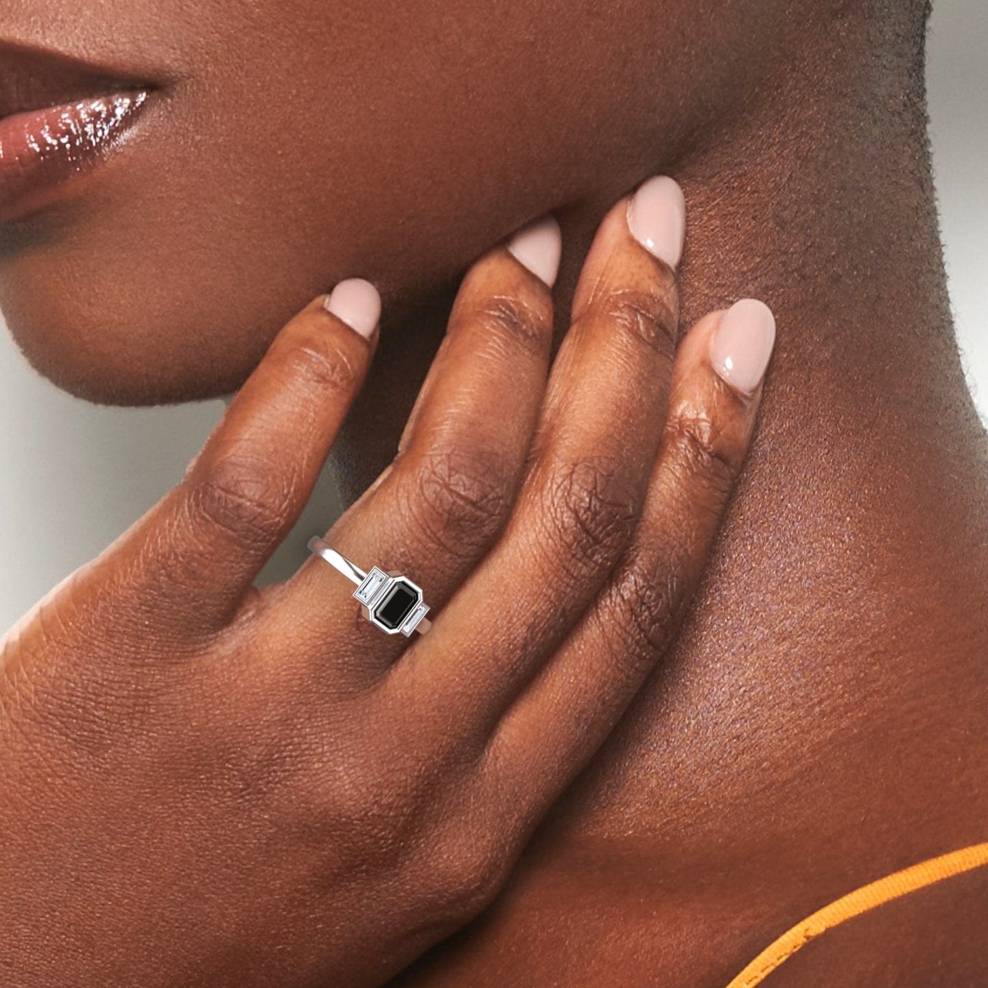 A hand with nude nails gently touches a smooth neck, displaying the Ivy Black & White Diamond Three-Stone Art Deco Ring—a striking silver piece with a large rectangular black gemstone—against a neutral background.