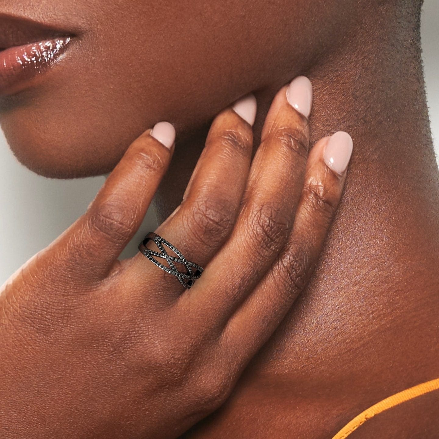 A close-up shows a hand with smooth brown skin and light pink nails gently touching the neck, highlighting the Black Diamond Criss-Cross Pave Band Ring - Eden on their finger.
