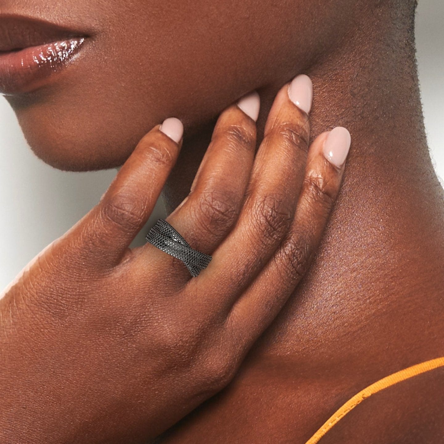 A close-up of a hand with smooth brown skin and pale pink nails gently touches the neck, showcasing the Black Diamond Pave Crossover Statement Ring - Noor. A thin orange strap is visible in the background.