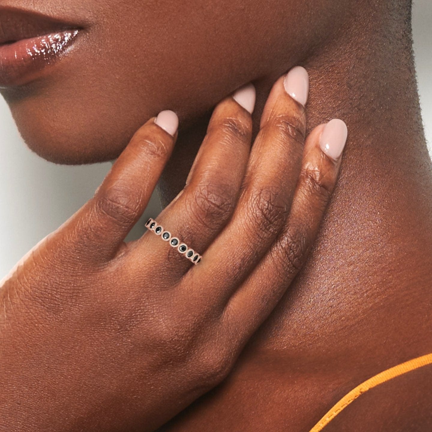 A close-up shows manicured fingers touching a smooth neck, wearing the Black Diamond Milgrain Bezel Set Eternity Band - Ira, a silver ring with black stones. An orange strap is visible on the persons shoulder.