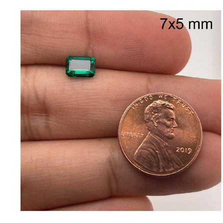 A hand displays a 7x5 mm 7x5 Exceptional Emerald Color Lab Created Emerald beside a 2019 U.S. penny for size comparison, with 7x5 mm text in the top right corner.