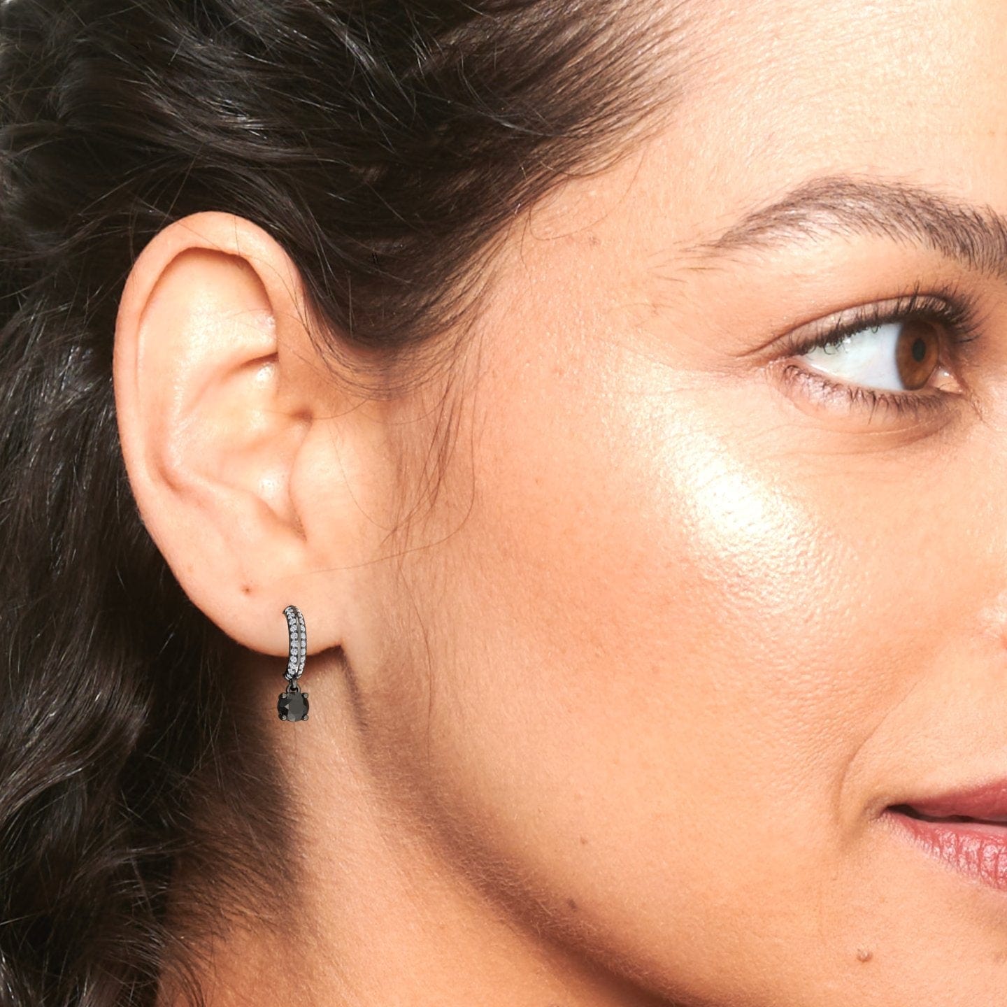 Close-up of a womans glowing face with dark hair pulled back, showcasing her ear adorned with the Serena Black & Diamond Pave Drop Hoop Earring.