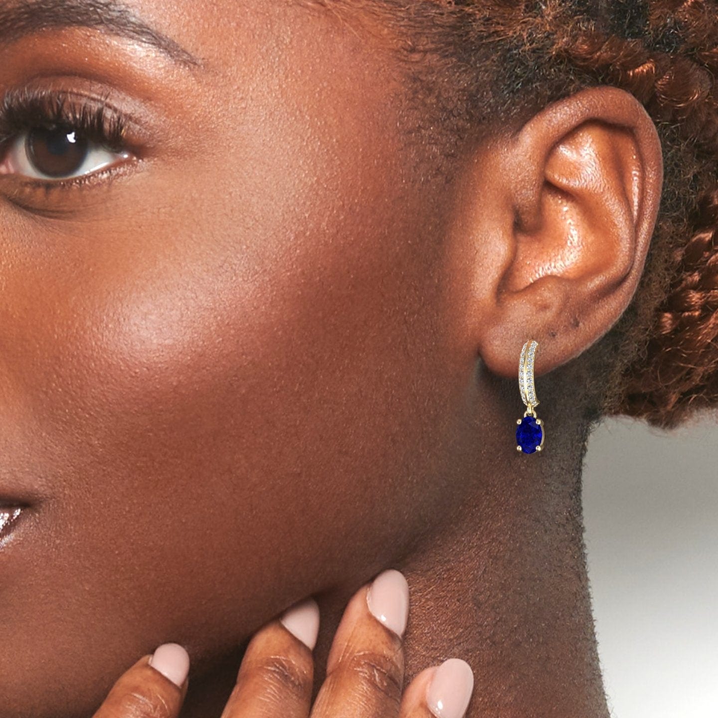Close-up of a woman wearing the Serena Blue Sapphire & Diamond Huggie Drop Earrings. Her manicured hand touches her chin, braided hair frames her ear, and the neutral background highlights the elegant jewelry.