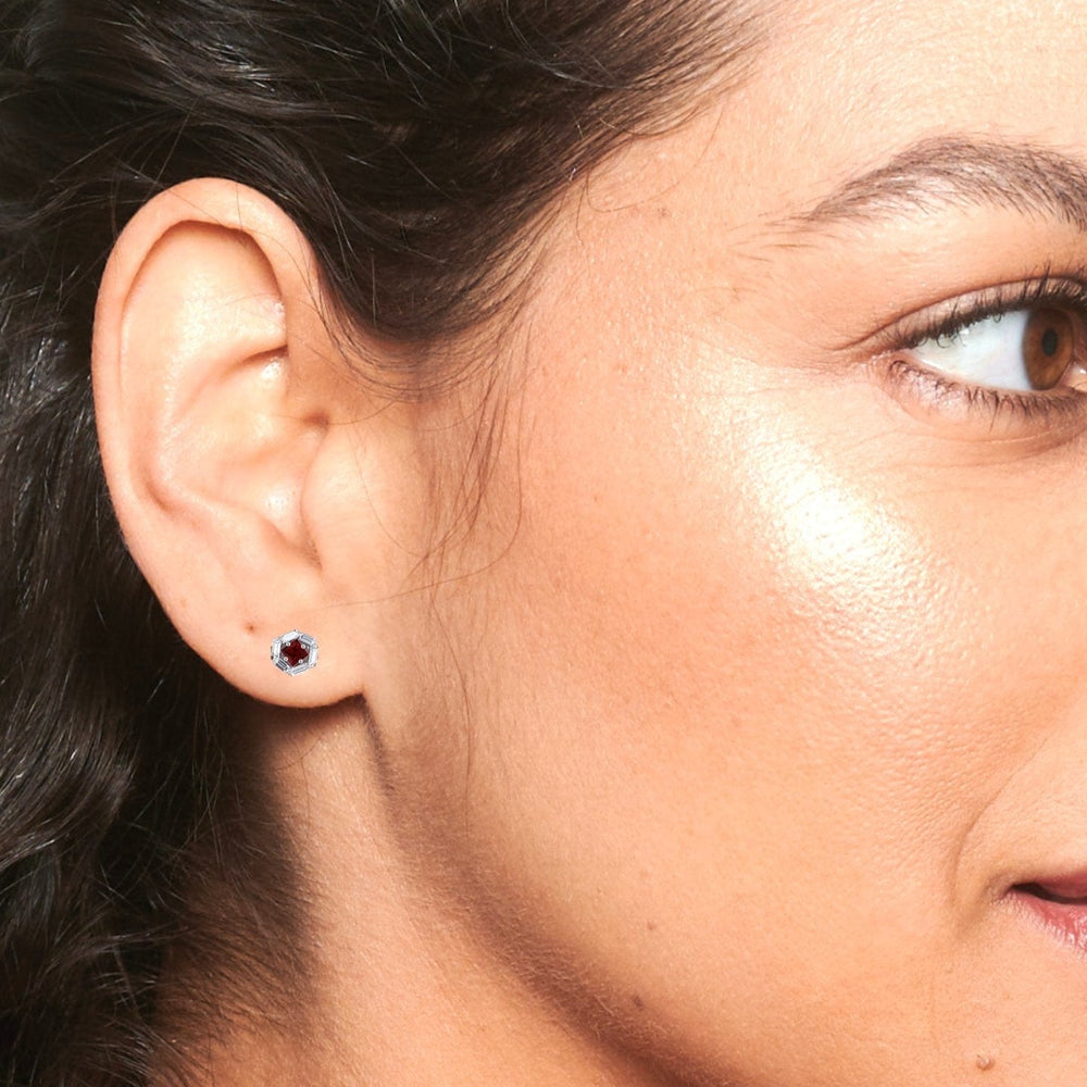 Close-up of a woman’s face focused on her ear, wearing the Ruby & Diamond Hexagon Halo Stud Earrings - Kiara. Her smooth skin, pulled-back hair, brown eye, and arched eyebrow frame the striking hexagon halo studs.