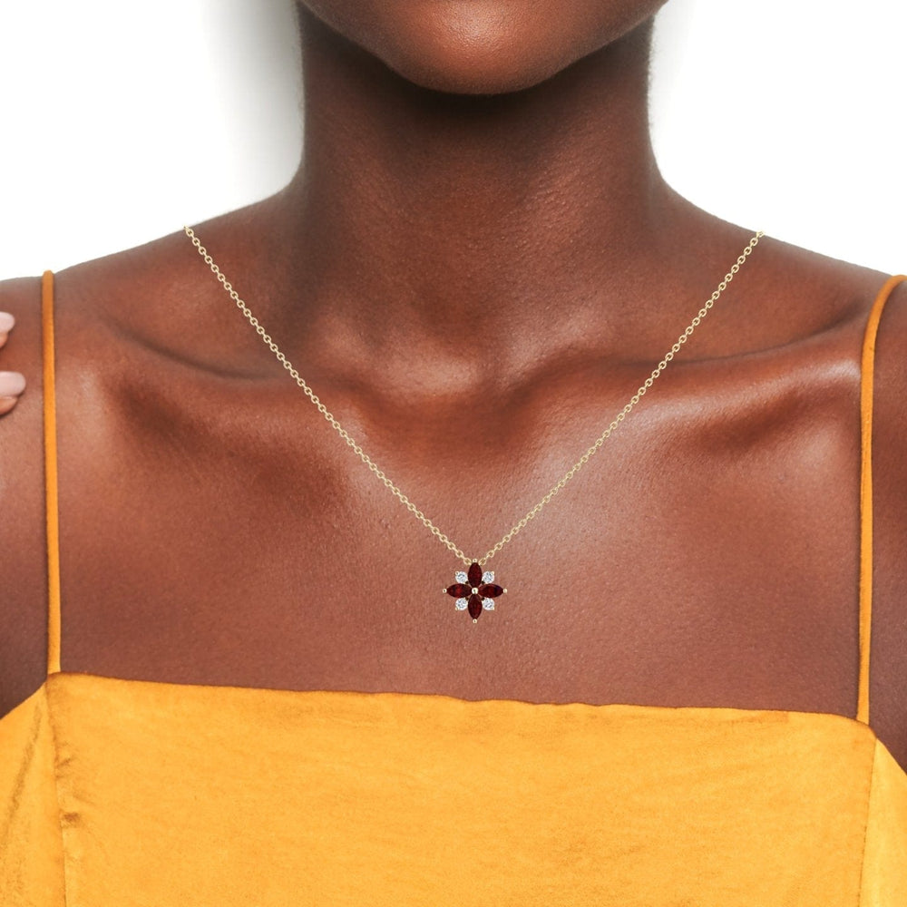 A woman in a yellow dress showcases the Ruby & Diamond Flower Necklace - Keira, featuring a red flower-shaped pendant, against a plain white background. Her shoulders and collarbone are visible.