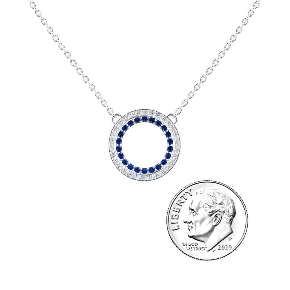 The Blue Sapphire & Diamond Double Halo Circle Necklace - Mona features a circular pendant with blue and clear stones, shown beside a U.S. dime for size reference.
