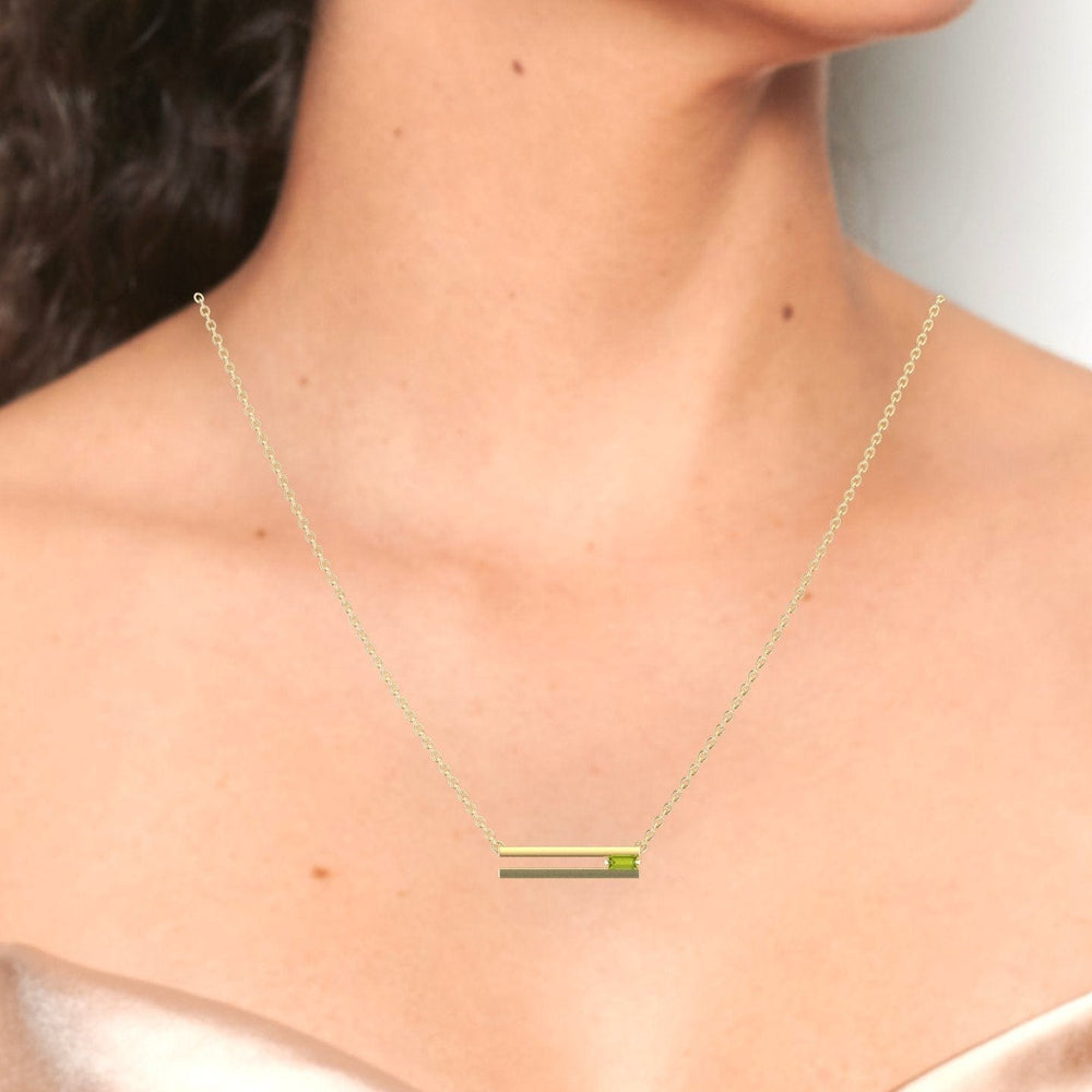 A woman in an off-shoulder, champagne-colored top showcases the Peridot Bar Necklace - Cecilia, featuring a delicate gold chain with a minimalist horizontal bar pendant at the center.