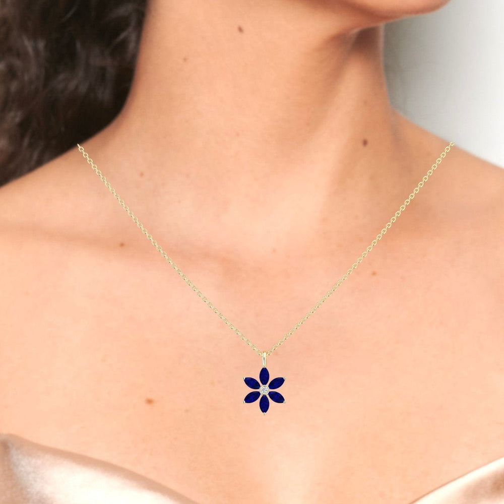 A woman shown from the shoulders up in an off-the-shoulder light top wears the Erin Lab Grown Blue Sapphire & Diamond Floral Necklace, featuring a delicate gold chain and a dark blue flower-shaped pendant.