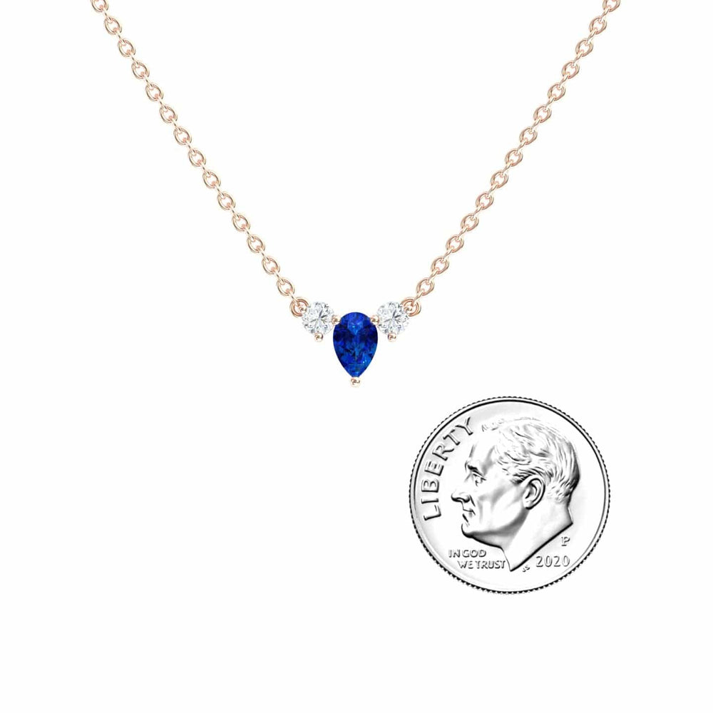 The Blue Sapphire & Diamond Three-Stone Necklace - Jina features a gold chain, a pear-shaped blue sapphire, and two round diamonds, shown next to a U.S. dime for size comparison.