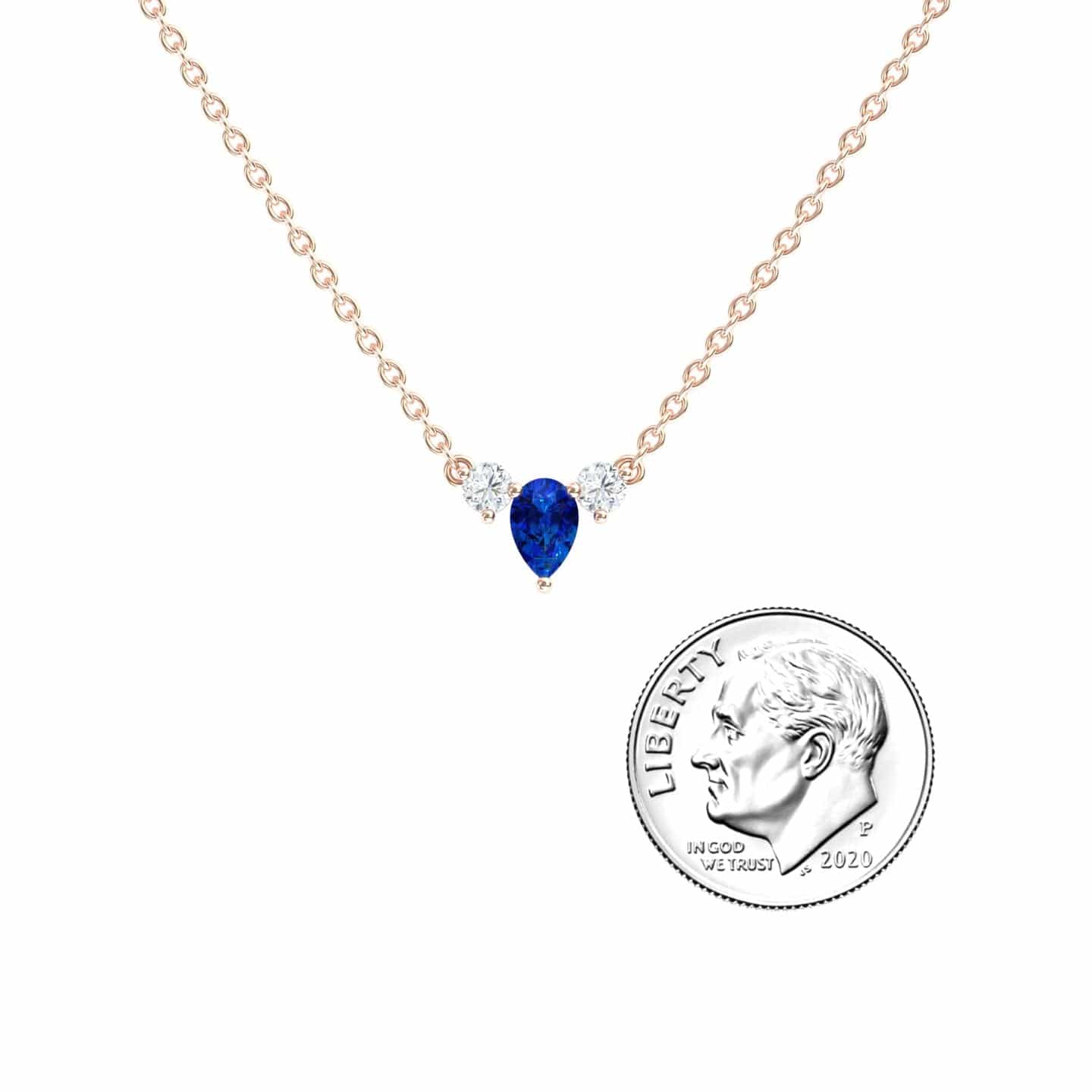 The Blue Sapphire & Diamond Three-Stone Necklace - Jina features a gold chain, a pear-shaped blue sapphire, and two round diamonds, shown next to a U.S. dime for size comparison.