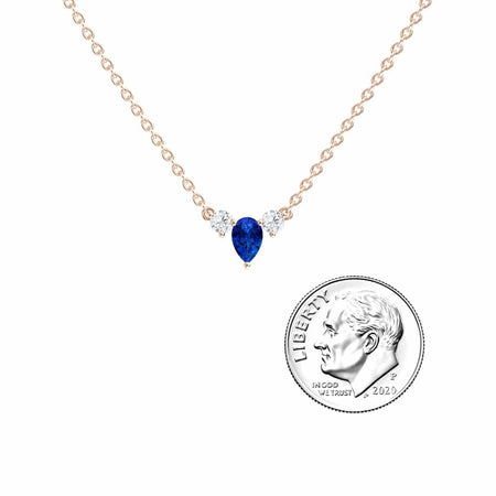 The Blue Sapphire & Diamond Three-Stone Necklace - Jina features a gold chain, a pear-shaped blue sapphire, and two round diamonds, shown next to a U.S. dime for size comparison.