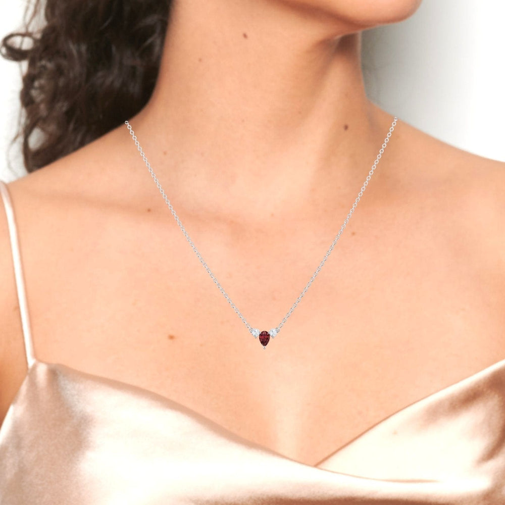 A woman in a champagne satin camisole showcases the Ruby & Diamond Three-Stone Necklace - Jina, featuring a delicate silver chain and a trio of dazzling stones. The focus is on her neck, collarbone, and the elegant jewelry.