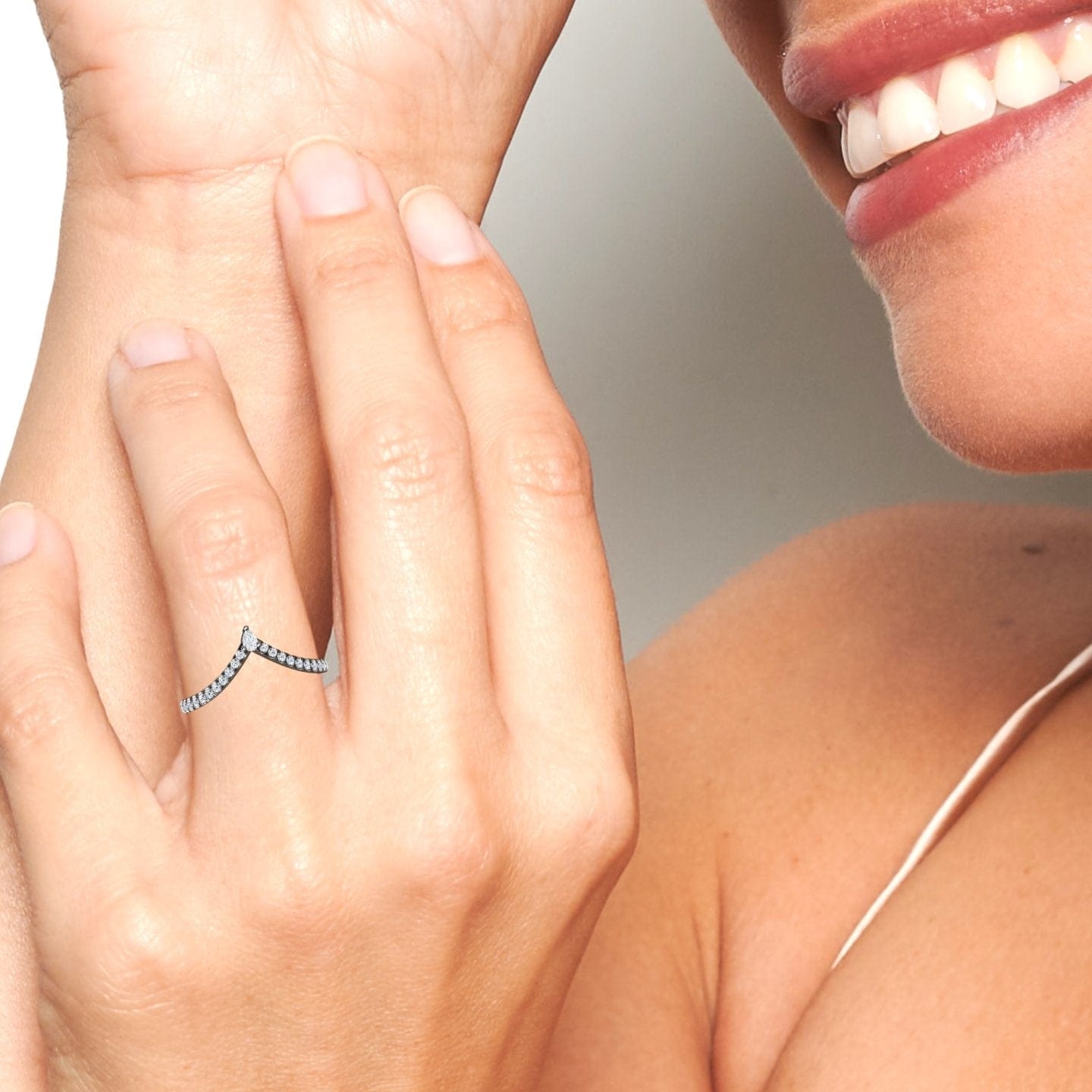 A close-up of a smiling person’s hand wearing the White Diamond Classic Chevron Wedding Band - Mei-Lin, a delicate V-shaped silver ring with small stones. Their face is partially visible, showing white teeth and smooth skin.